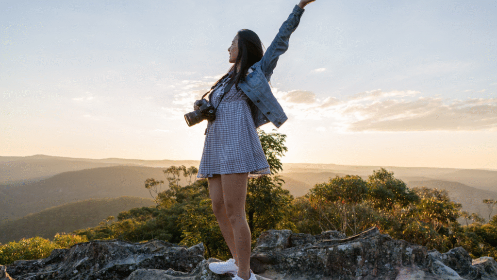 Mujer con cámara levantando los brazos en un paisaje natural, símbolo de felicidad y consciencia plena.